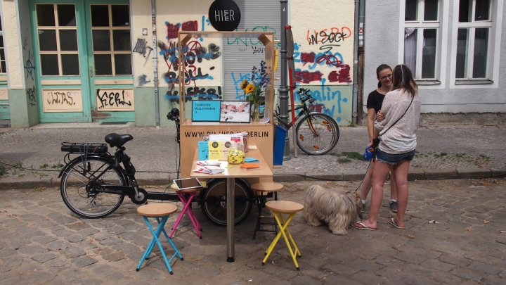 Das Lastenrad von "Hier in Berlin" ist als Infostand aufgebaut. Zwei junge Frauen mit Hund unterhalten sich.