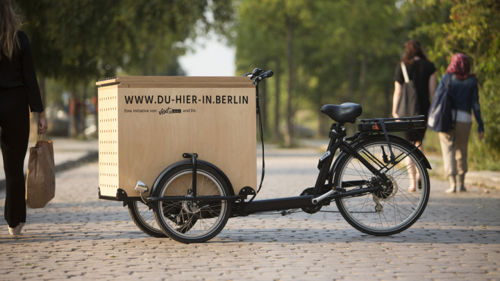 Das Lastenrad mit der Beschriftung "www.du-hier-in.berlin - Eine Initiative von visitBerlin und dir" steht auf einer Straße.