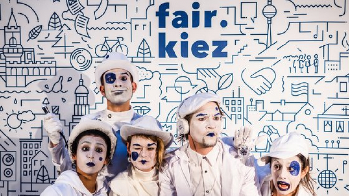 Pantomime-Künstler:innen und Kommunikator:innen stehen vor einer weißen Leinwand mit dem blauem Schriftzug "fair.kiez.". Sie sind weiß gekleidet und geschminkt, mit blauen Akzenten.