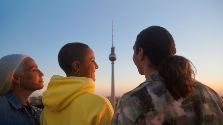 Drei junge Personen schauen fröhlich zum Horizont. Zwischen ihnen ist der Berliner Fernsehturm zu sehen.